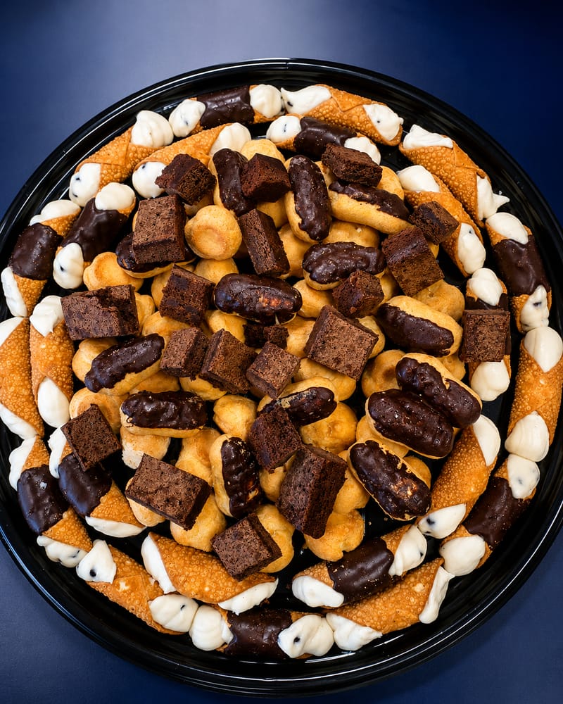 Desserts on a black round tray, seen from above: chocolate-dipped cannoli piped with cream around the rim, chocolate eclairs, brownie squares and cream puffs heaped in the middle